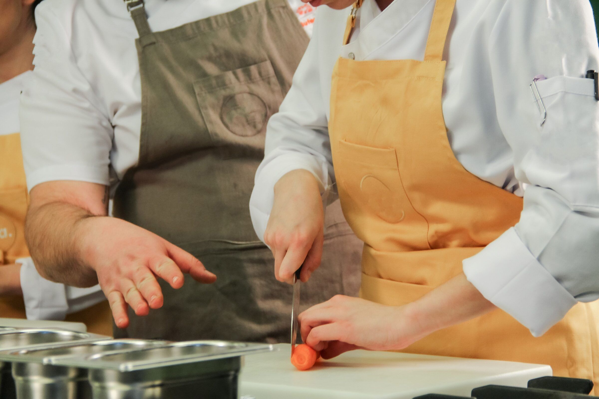 Close-up of a chef's hands cutting a carrot on a cutting board while a colleague looks on.